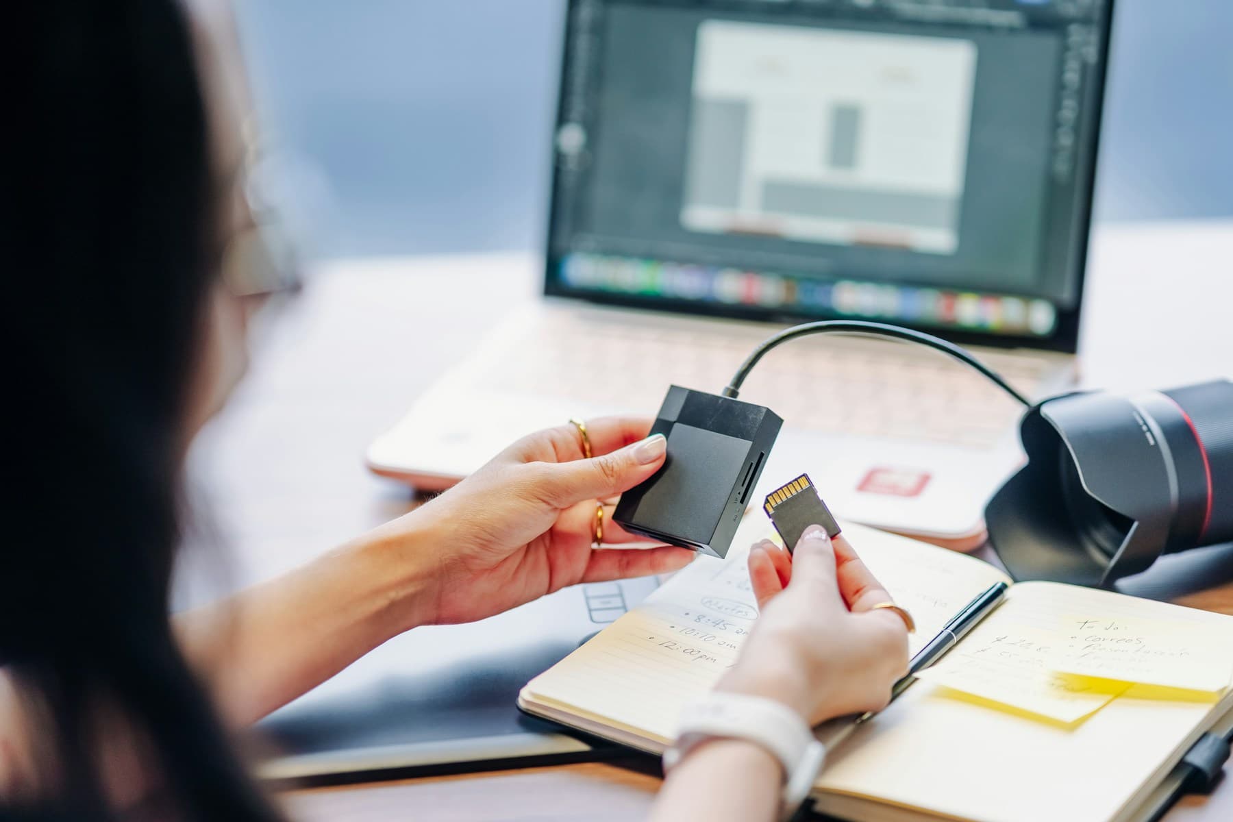 A person holding a memory card beside a laptop and camera lens